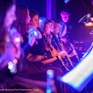 Koncert muzyki filmowej - Crushed Sounds BigBand - Grupa ludzi siedzi na krzesłach i gra na klarnetach patrząc w nuty, pada na nich różowe światło. - Fot. Przemysław Sroka