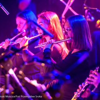 Koncert muzyki filmowej - Crushed Sounds BigBand - Grupa kobiet siedzi na krzesłach i gra na fletach patrząc w nuty, pada na nie różowe światło.  - Fot. Przemysław Sroka
