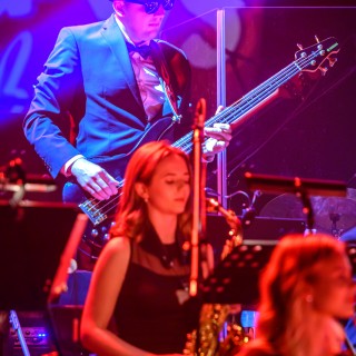 Koncert muzyki filmowej - Crushed Sounds BigBand - Mężczyzna w kapeluszu i okularach gra na gitarze, przed nim kobieta siedzi na krześle i w rękach trzyma puzon.  - Fot. Przemysław Sroka
