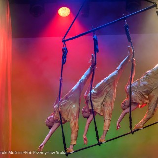 "Four Seasons" - Akrobatyczny Teatr Tańca Mira-Art. Scena Otwarta 2021 - Trzy kobiety stoją na czarnym wiszącym przyrządzie akrobatycznym, schylają się w dół, stojąc na jednej nodze. Oświetlają je kolorowe światła. - Fot. Przemysław Sroka