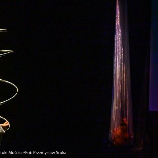 "Four Seasons" - Akrobatyczny Teatr Tańca Mira-Art. Scena Otwarta 2021 - Artyści przedstawiają występ "Four Seasons". Artystka z lewej strony kładzie się na końcu metalowego rekwizytu w postaci metalowej spirali. Po prawej stronie dwie osoby znajdują się w wiszących, przezroczystych foliach.  - Fot. Przemysław Sroka