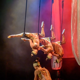 "Four Seasons" - Akrobatyczny Teatr Tańca Mira-Art. Scena Otwarta 2021 - Trzy artystki wiszą w artystycznej figurze na akrobatycznym przyrządzie z głową skierowaną w dół. Przed nimi znajduje się kobieta, klęcząca tyłem na scenie . - Fot. Przemysław Sroka