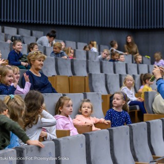 „O trzech takich, od których wszystko się zaczęło" - koncert Filharmonii Krakowskiej z cyklu "Bajki muzyką pisane" - Grupa dzieci siedzi na miejscach na sali widowiskowej. Z prawej strony opiekunka stoi przed nimi i robi im zdjęcie. - Fot. Przemysław Sroka