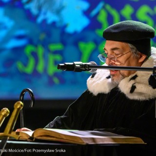„O trzech takich, od których wszystko się zaczęło" - koncert Filharmonii Krakowskiej z cyklu "Bajki muzyką pisane" - Starszy mężczyzna w czarnym płaszczu z ozdobnym, futrzanym kołnierzem, w czapce oraz okularach czyta księgę lężącą na stole. - Fot. Przemysław Sroka