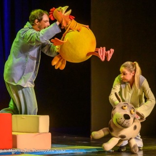 Teatr Młodych Widzów - "Miś Tymoteusz" - spektakl Teatru Rabcio - Na scenie po lewej stronie za dużymi kolorowymi klockami stoi mężczyzna unoszący dużą, pluszową kurę. Naprzeciwko niego klęczy kobieta trzymająca dużego, zabawkowego psa.  - Fot. Przemysław Sroka