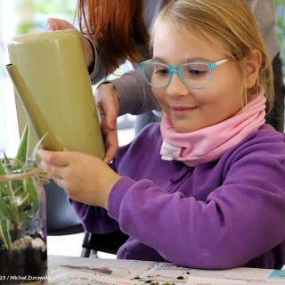 „Ogród w słoiku” - warsztaty - Kobieta podaje dziecku konewkę. Dziewczynka patrzy na roślinę w słoiku i uśmiecha się. - fot. Michał Żurowski