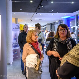 "Women" - wystawa fotografii Doroty Góreckiej - Trzy kobiety stoją obok siebie w holu. Jedna z nich coś mówi, druga patrzy na nią i się uśmiecha. Trzecia patrzy na zdjęcie przedstawiające kobietę o długich włosach. - Fot: Przemysław Sroka
