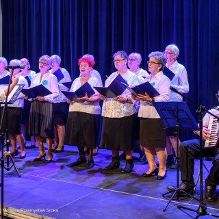 Senior w Centrum - koncert chóru Siemaszki - Kobiety trzymają śpiewniki w dłoniach i śpiewają. Każda z nich jest ubrana w białą bluzkę, do której przypięty jest niebieski kotylion. Przed nimi stoi dyrygentka, a po prawej stronie siedzi mężczyzna grający na akordeonie. - Fot: Przemysław Sroka