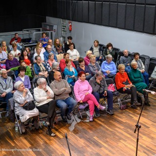 Senior w Centrum - koncert chóru Siemaszki - Publiczność siedzi na krzesłach. Przed nimi stoi kobieta z mikrofonem w dłoni. - Fot: Przemysław Sroka