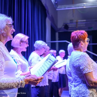 Senior w Centrum - koncert chóru Siemaszki - Kobiety ubrane w białe bluzki stoją na scenie, trzymają śpiewniki w dłoniach i śpiewają. - Fot: Przemysław Sroka