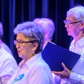 Senior w Centrum - koncert chóru Siemaszki - Kobiety trzymają śpiewniki w dłoniach i śpiewają. Każda z nich jest ubrana w białą bluzkę, do której przypięty jest niebieski kotylion. - Fot: Przemysław Sroka