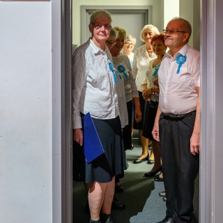 Senior w Centrum - koncert chóru Siemaszki - Kobiety i mężczyzna stoją w garderobie. Wszyscy mają niebieskie kotyliony przypięte do ubrań. - Fot: Przemysław Sroka