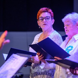 Senior w Centrum - koncert chóru Siemaszki - Kobiety trzymają śpiewniki w dłoniach i śpiewają. Każda z nich jest ubrana w białą bluzkę, do której przypięty jest niebieski kotylion. - Fot: Przemysław Sroka