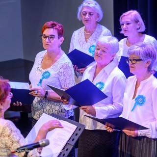 Senior w Centrum - koncert chóru Siemaszki - Kobiety trzymają śpiewniki w dłoniach i śpiewają. Każda z nich jest ubrana w białą bluzkę, do której przypięty jest niebieski kotylion. Przed nimi stoi dyrygentka. - Fot: Przemysław Sroka