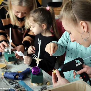 „Stroiki bożonarodzeniowe” warsztaty dla grup zorganizowanych przedszkolnych i wczesnoszkolnych - Dwie kobiety pomagają dzieciom przyklejać ozdoby do gąbki florystycznej. - fot. Michał Żurowski