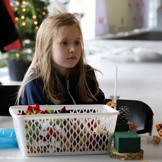 „Stroiki bożonarodzeniowe” warsztaty dla grup zorganizowanych przedszkolnych i wczesnoszkolnych - Dziewczynka siedzi przy stole. Przed nią leży koszyk koszyk z kokardkami. - fot. Michał Żurowski